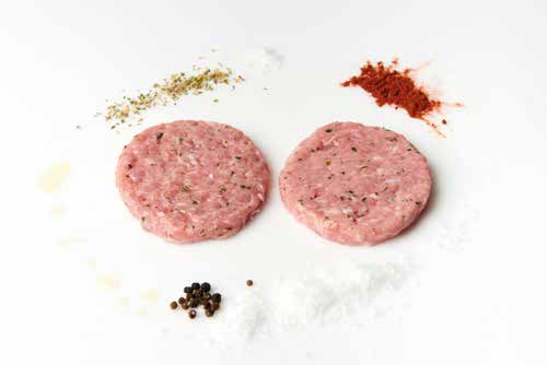 Hamburguesas de conejo sobre una superficie blanca rodeadas por las especias que contiene: granos de pimienta, sal, pimentón, y demás especias.